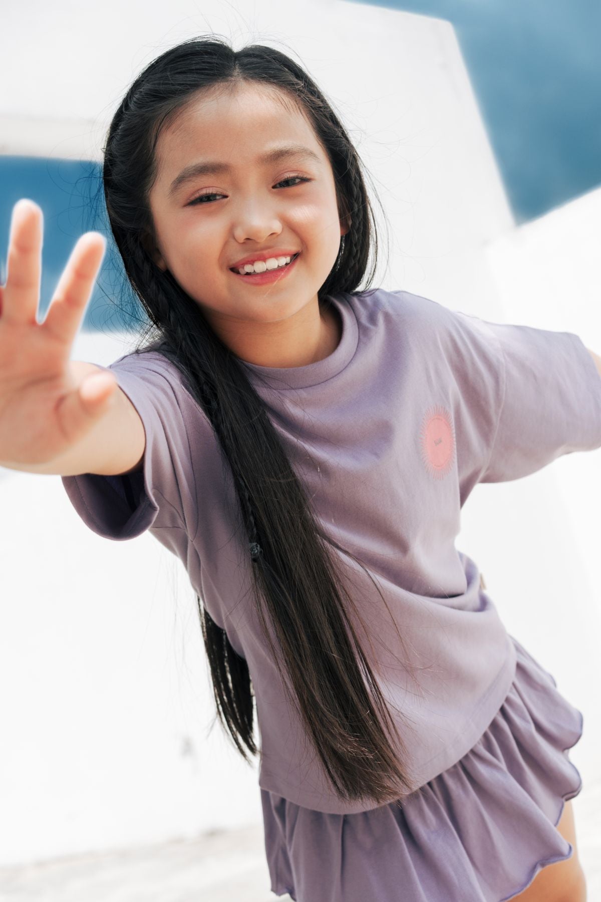 Young girl in a lavender ruffle skirt and matching t-shirt, smiling confidently with her hand reaching toward the camera