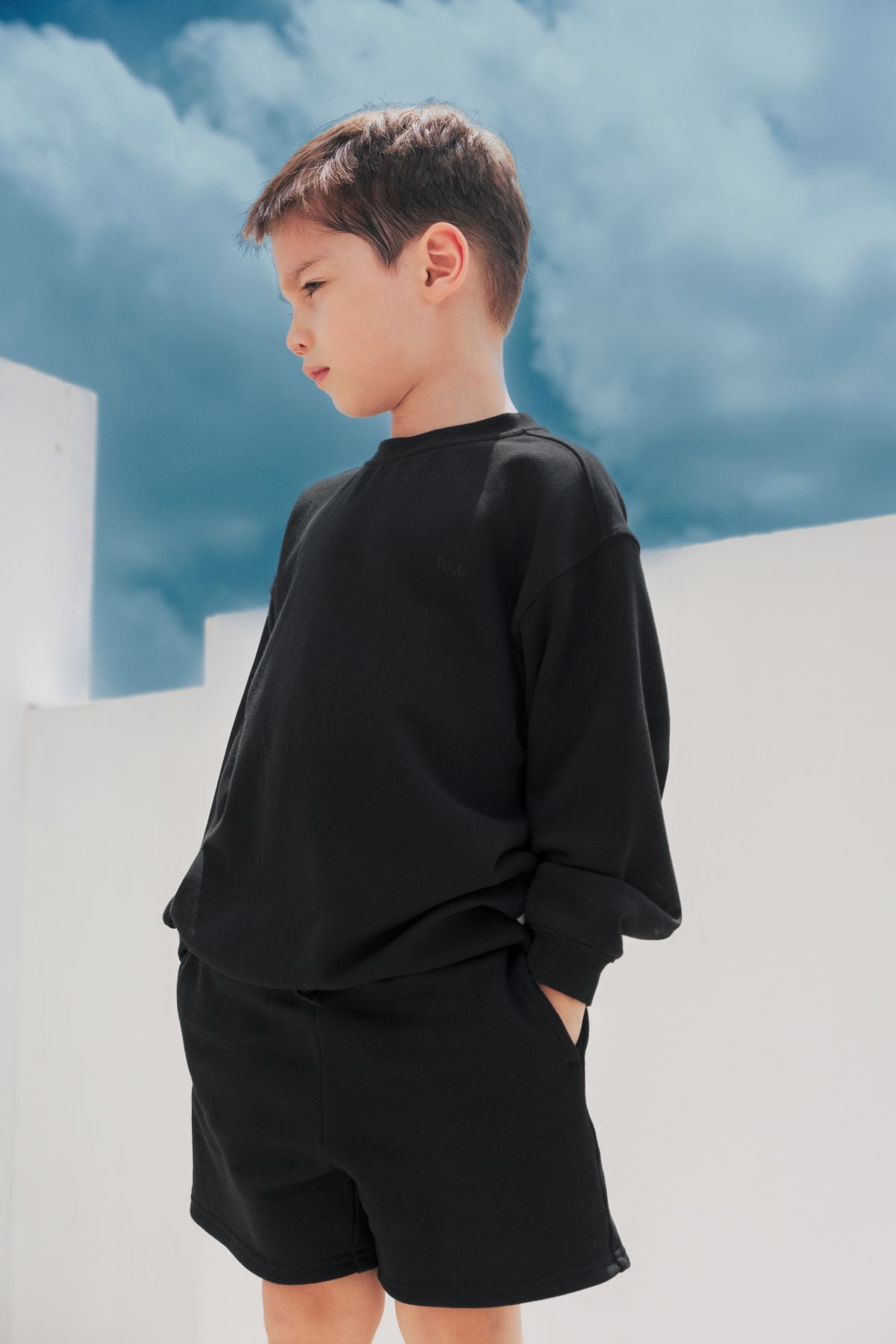 Confident child in a black denim kidswear set sitting on stairs under natural light, showcasing a modern minimalist outfit.