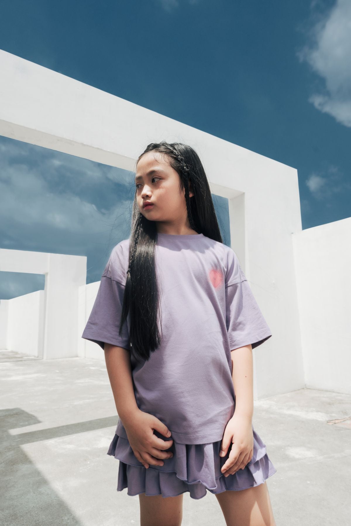 Young girl in a lavender oversized t-shirt and ruffle skirt standing confidently under a bright sky with modern minimalist architecture in the background.